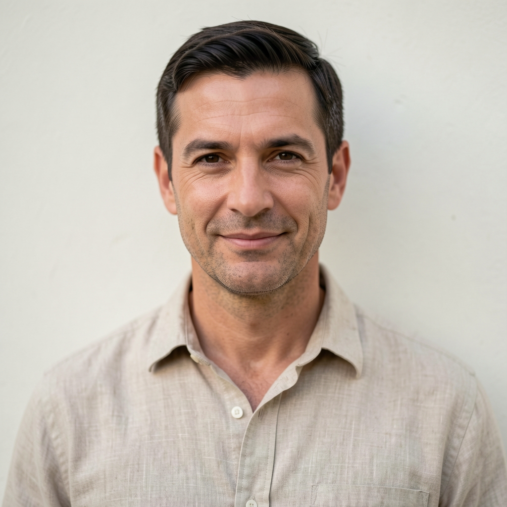 Professional headshot of a man in his early 40s with short dark hair, wearing a natural linen shirt, standing against a plain off-white wall in soft diffused light
